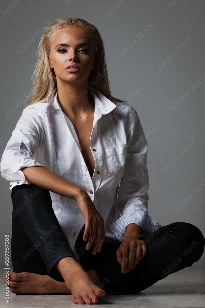Portrait of blond young woman with beautiful makeup and hairstyle in white shirt sits on gray background. Fashion model poses in elegant clothes in studio Portrait of blond young woman with beautiful makeup and hairstyle in white shirt sits on gray background. Fashion model poses in elegant clothes in studio