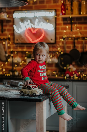 Portrait of little blond boy in red and green Christmas pyjamas sitting on a kitchen table playing with retro car against lights.