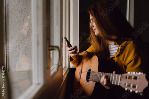 Smiling redheaded woman with guitar sitting at open window looking at cell phone photo