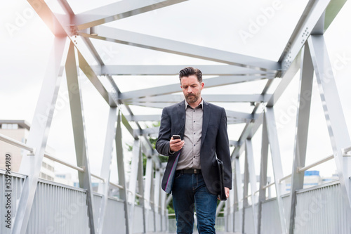 Wallpaper Mural Confident handsome businessman using smart phone while walking on footbridge in city Torontodigital.ca