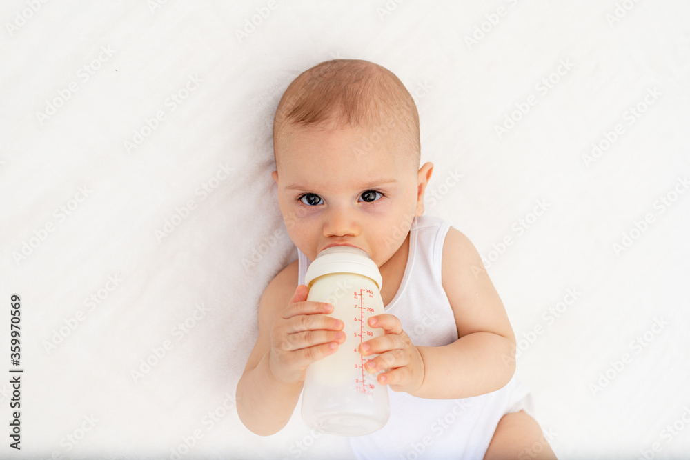 baby boy 8 months old lies drinking milk from a bottle on the bed in the nursery, feeding the baby, baby food concept baby boy 8 months old lies drinking milk from a bottle on the bed in the nursery, feeding the baby, baby food concept