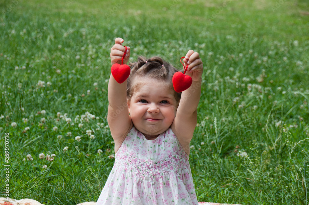 Very beautiful baby girl sitting on lawn in a park and plays cheerfully, holds red decorative hearts in her hands Very beautiful baby girl sitting on lawn in a park and plays cheerfully, holds red decorative hearts in her hands