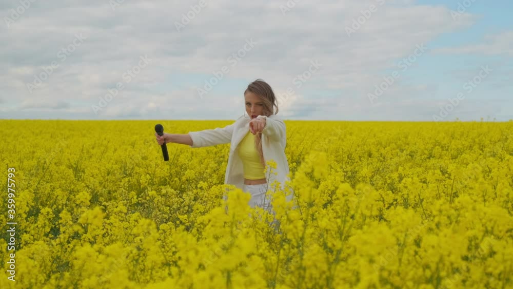 An energetic female singer with a microphone in a field of rapeseed with yellow flowers jumps shakes her head and dances An energetic female singer with a microphone in a field of rapeseed with yellow flowers jumps shakes her head and dances