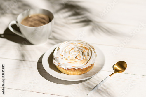 Fototapeta Naklejka Na Ścianę i Meble -  Lemon meringue little pie on a wooden board, table on a palm beach outdoor cafe in the shadows of palm branches. Continental breakfast with a cup of coffee.