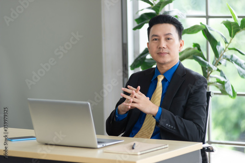 Portrait of middle age Asian businessman wearing formal black suit sitting on chair, there are laptop and smartphone at his desk, looking at camera, working in workplace