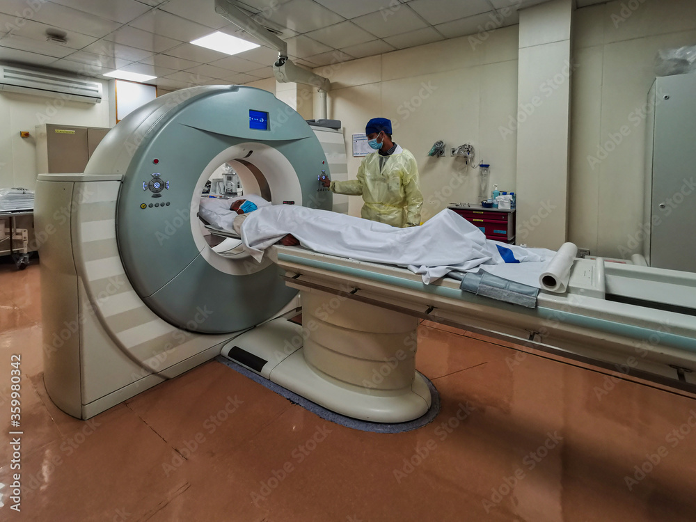 Computed Tomography machine (CT) in Radiology Department with radiology technician wearing personal protective equipment (PPE) preparing COVID-19 patient for examination Computed Tomography machine (CT) in Radiology Department with radiology technician wearing personal protective equipment (PPE) preparing COVID-19 patient for examination