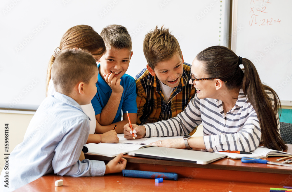 Female teacher helps school kids to finish they lesson.They sitting all together at one desk. Female teacher helps school kids to finish they lesson.They sitting all together at one desk.