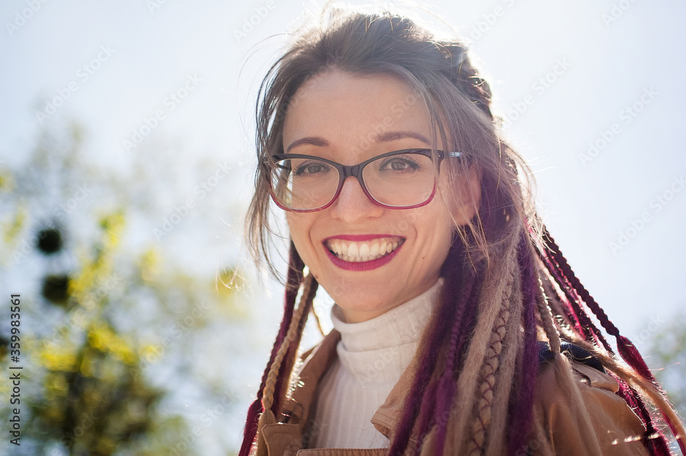 Young girl with long dreadlocks and eyeglasses in nude color trench is looking at the camera with charming smile on sky background. Positive psychology concept
