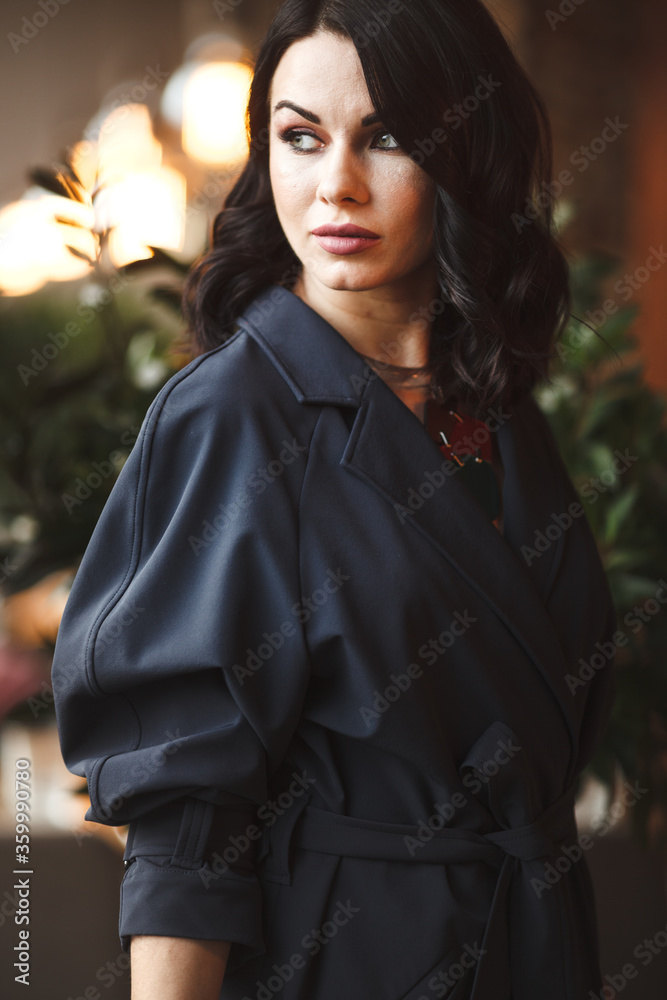 Fototapeta premium Lifestyle and citylife. Beautiful brunette business lady in modern gems and fashionable navy coat having a lunch in cafe. Woman sits by the table and holds cup of tea