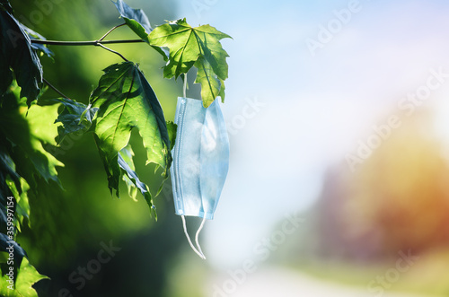 A medical mask hangs on a branch of a green tree. The period after quarantine in nature. Sunny.