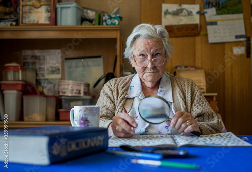 Old lady at dining table doing crossword puzzle with dictionary, glasses, magnifying glass, tea photo