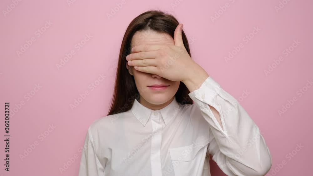 Young girl covers her face with hand, opens mouth, tries to hide from someone, female being shocked, covers eyes waits for surprise, dressed in white shirt, isolated over pink studio background Young girl covers her face with hand, opens mouth, tries to hide from someone, female being shocked, covers eyes waits for surprise, dressed in white shirt, isolated over pink studio background