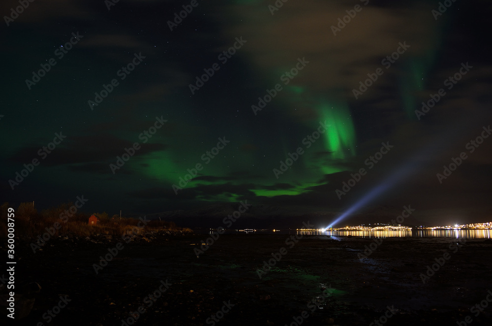 aurora borealis over a boat searchlight on the fjord reflecting of the surface with illuminated settlement in the background