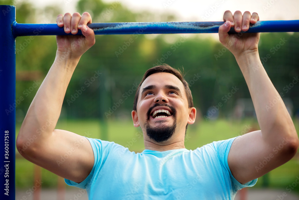 Naklejka premium Cheerful middle-aged man doing pull ups exercise on shabby steel bar outdoors in summer evening. Tanned bearded man in blue shirt doing workout at training ground. Strength and motivation concept