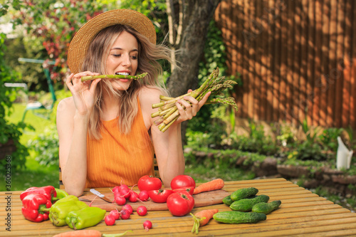A young pretty girl in a hat is sitting at a wooden table with fresh vegetables grown in her garden in the summer.