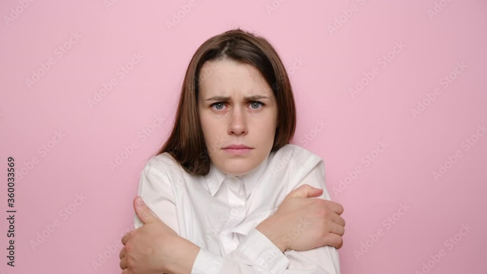 Young woman freezes in light clothing warms up by hugging herself, seasonal changes low temperature heating problem, looks at camera, chills feeling of coldness concept, isolated on pink background Young woman freezes in light clothing warms up by hugging herself, seasonal changes low temperature heating problem, looks at camera, chills feeling of coldness concept, isolated on pink background
