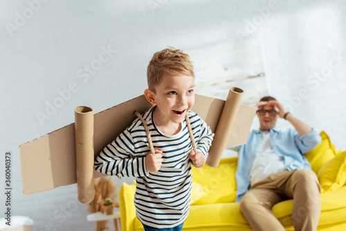 selective focus of happy boy with carton plane wings on back imitating flying near father