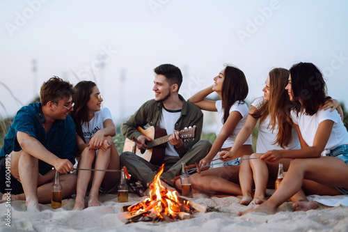 Group of happy friends frying sausages on campfire at the beach. A company of young people came together for a barbecue.  © maxbelchenko