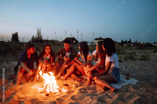 Group of happy friends frying sausages on campfire at the beach. A company of young people came together for a barbecue. 