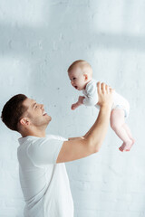 cheerful young father holding cute infant in outstretched hands in bedroom