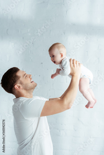 cheerful young father holding cute infant in outstretched hands in bedroom