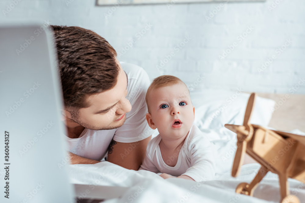 Obraz premium selective focus of young father looking at surprised baby boy while lying in bed near laptop and toy plane