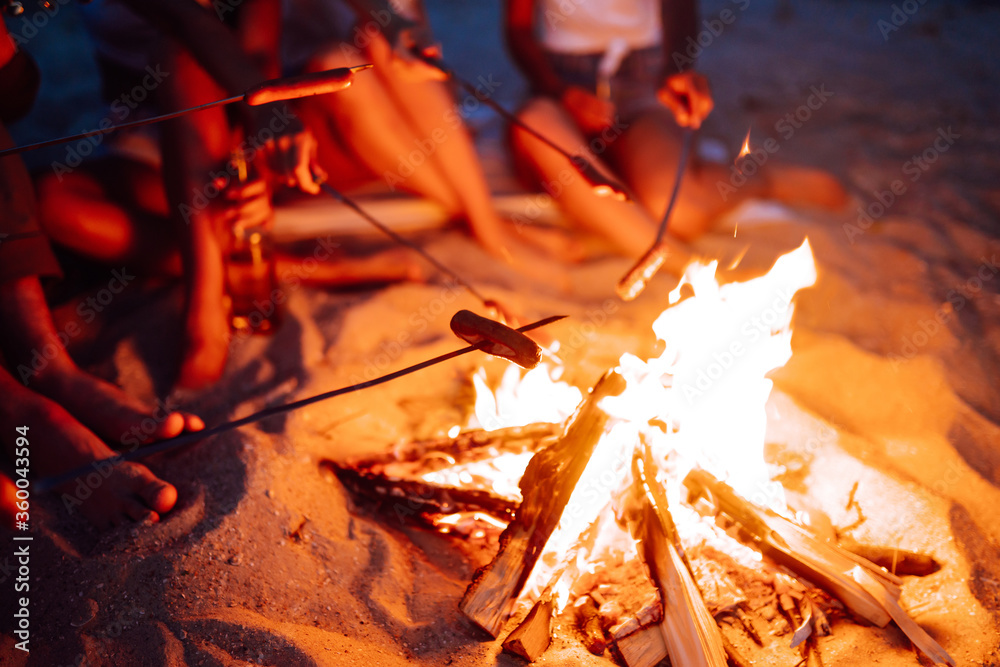 Travel, tourism, hike, picnic and people concept - group of happy friends frying sausages on campfire at the beach. A company of young people came together for a barbecue.