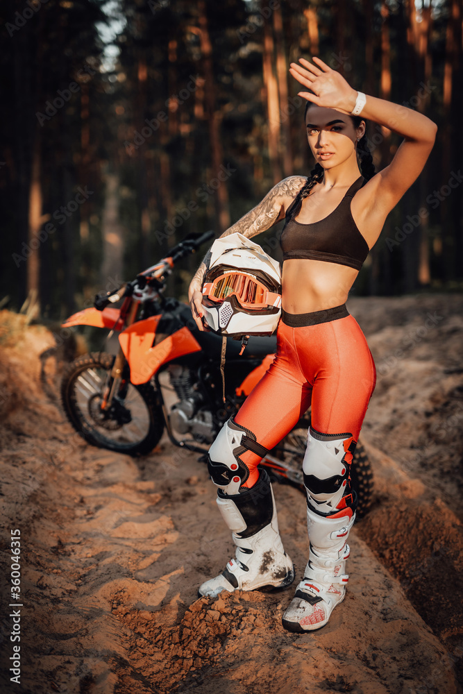 Obraz premium Beautiful brunette girl wearing motocross outfit with semi naked torso standing on a sand trail in the forest, holds his helmet and covers his eyes with his hand from the bright sun