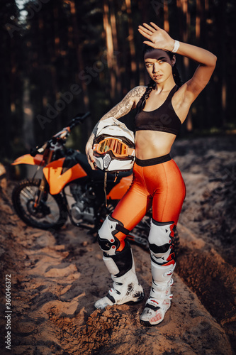 Beautiful brunette girl wearing motocross outfit with semi naked torso standing on a sand trail in the forest  holds his helmet and covers his eyes with his hand from the bright sun