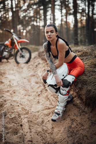 Beautiful brunette girl wearing motocross outfit with semi naked torso sitting on a grass in the forest