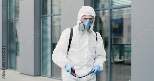 Caucasian man in special antibacterial white suit and respirator walking the street and disinfecting sidewalk with pulverizator. Spraying disinfectant on ground outdoors. Coronavirus pandemic concept. photo