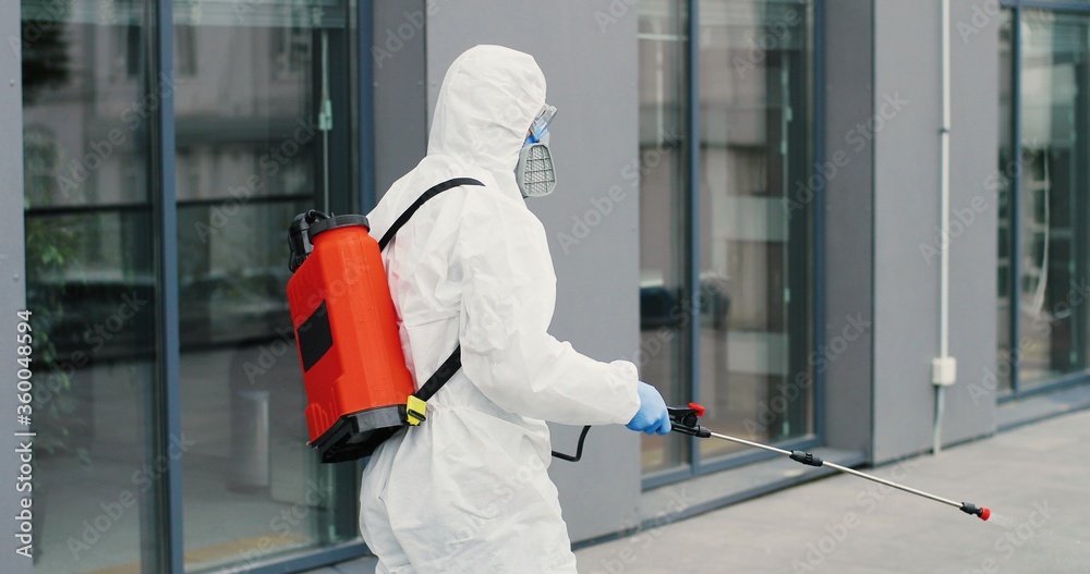 Caucasian man in special antibacterial white suit and respirator walking the street and disinfecting sidewalk with pulverizator. Spraying disinfectant on ground outdoors. Coronavirus pandemic concept.