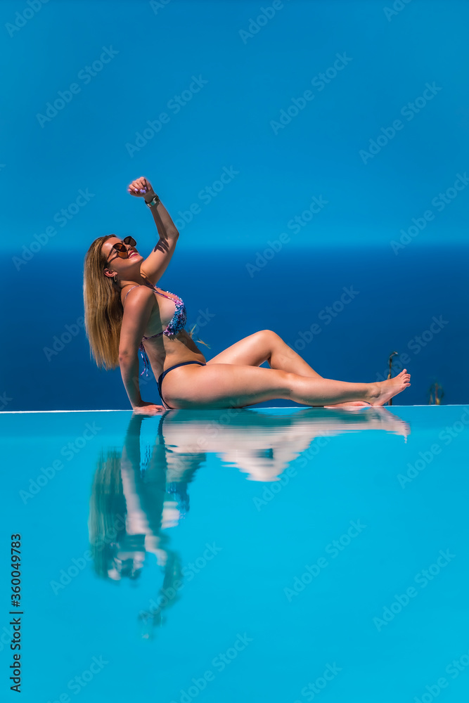 Summer lifestyle in an infinity pool with a young blonde Caucasian woman in a pink and purple bikini with sunglasses. Enjoying the pool reflected in the water