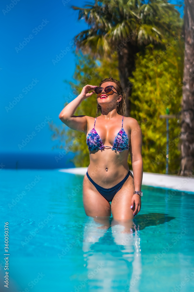 Summer lifestyle in an infinity pool with a young blonde Caucasian woman in a pink and purple bikini with sunglasses. Enjoying the pool on a summer afternoon