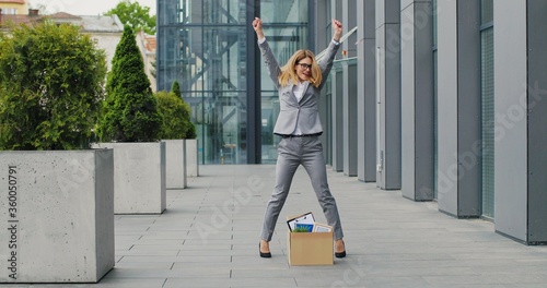 Caucasian happy businesswoman walking the street with box of stuff as leaving business center and dancing. Female office worker leaving job and celebrating. Fired cheerful woman.