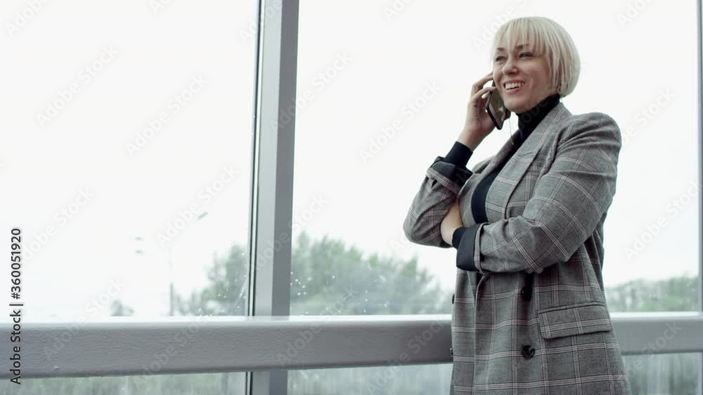 A side view slow motion close up shot of a young blonde business woman standing by the window inside an office building and having a conversation with her colleague on the phone A side view slow motion close up shot of a young blonde business woman standing by the window inside an office building and having a conversation with her colleague on the phone