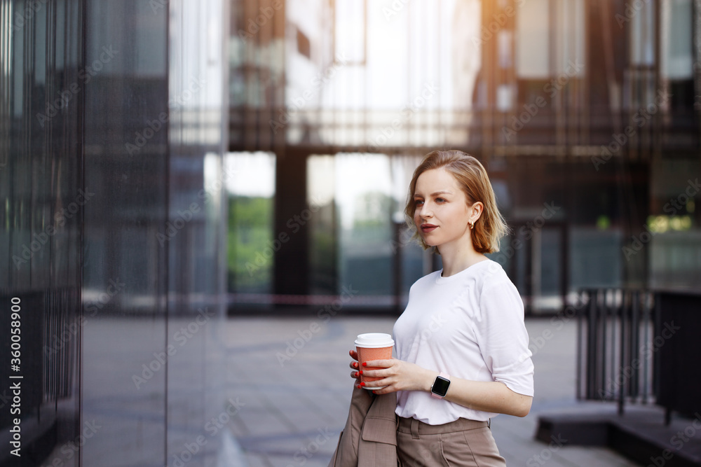 Business woman in beige suit, holding cup of coffee, walking outdoors on city street with skyscraper or building background, looking on window. Break at work, leisure time, enjoyment, shopping