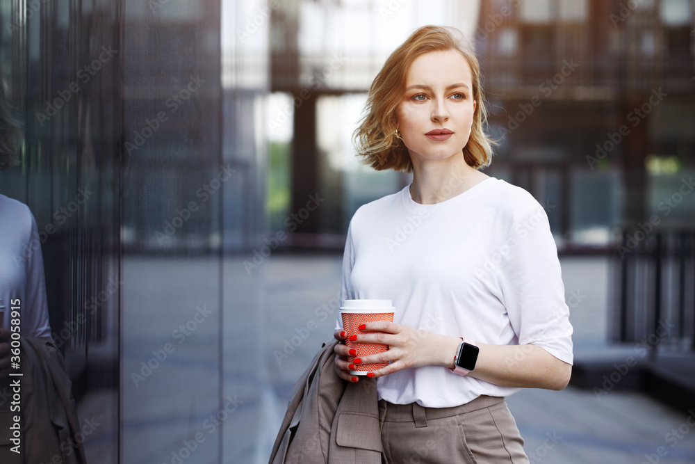 Business woman in beige suit, holding cup of coffee, walking outdoors on city street with skyscraper or building background. Break at work, leisure time, enjoyment, shopping near window
