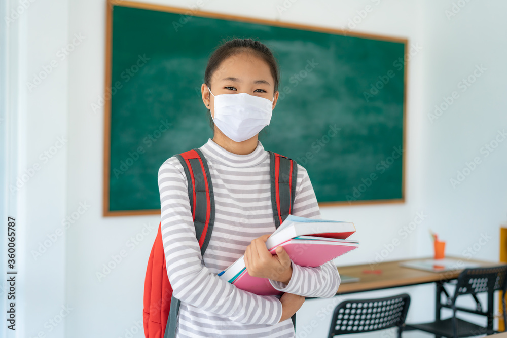 Asian primary students girl with backpack and books wearing masks to prevent the outbreak of Covid 19 in classroom while back to school reopen their school, New normal for education concept..