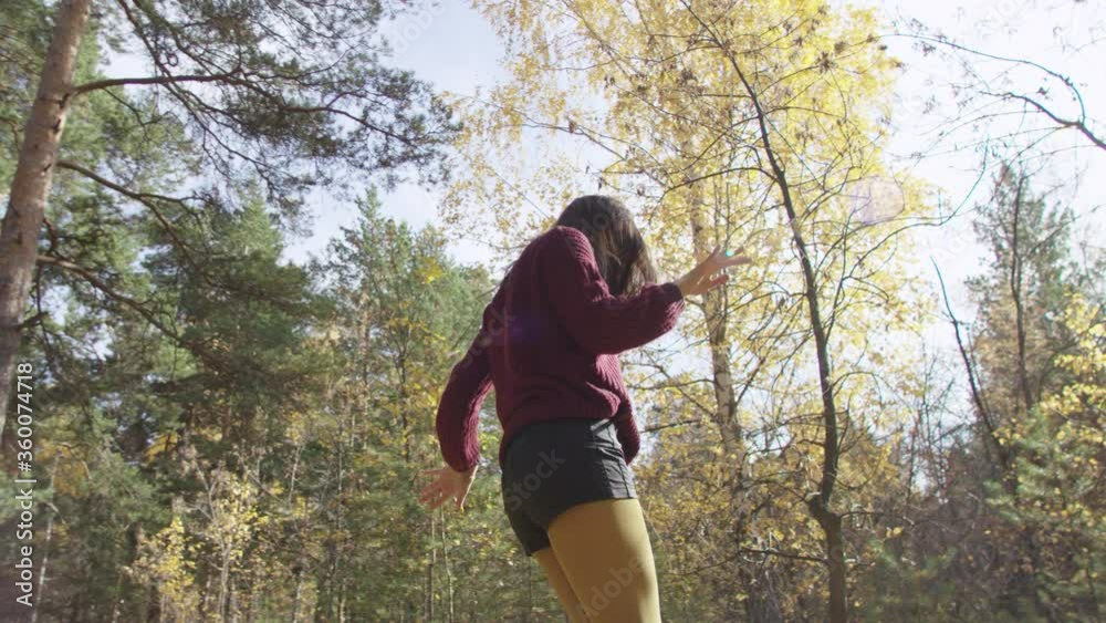 4K low angle portrait of female dressed in red jumper, black shorts and yellow tights dancing happily in the middle of autumn forest. 360 degree tracking arc shot.