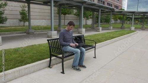 High quality 4k video of a young men who is sitting on a train station and working on his laptop which is holding on the lap. Young men sitting on a black metal banch on a station in the city. 