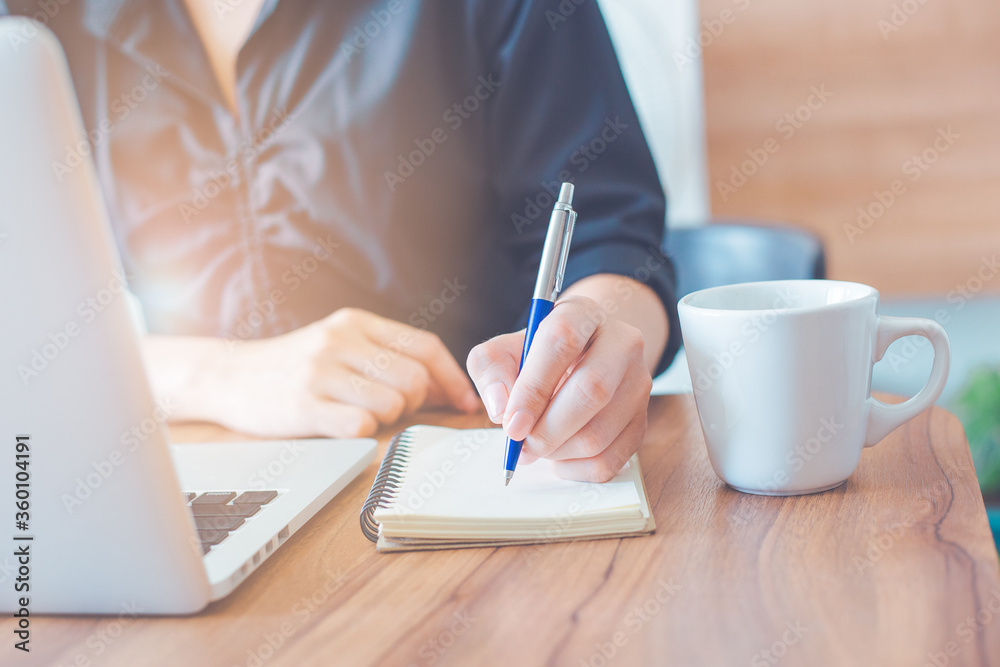 Business woman is writing on a notebook with a pen and using a laptop to work in the office.