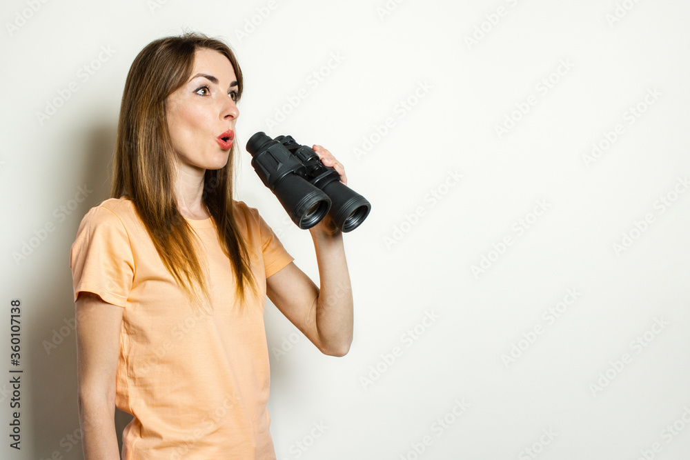 Young friendly girl with a surprised face in a T-shirt is holding binoculars on a light background. Banner. Emotional face Young friendly girl with a surprised face in a T-shirt is holding binoculars on a light background. Banner. Emotional face