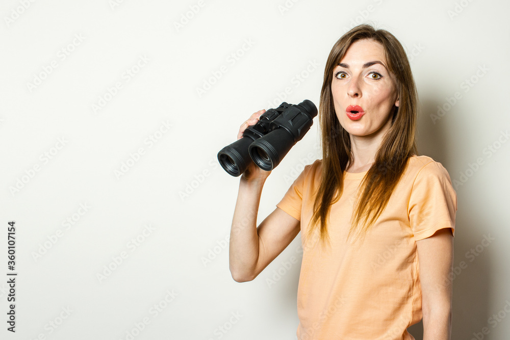 Young friendly girl with a surprised face in a T-shirt is holding binoculars on a light background. Banner. Emotional face Young friendly girl with a surprised face in a T-shirt is holding binoculars on a light background. Banner. Emotional face