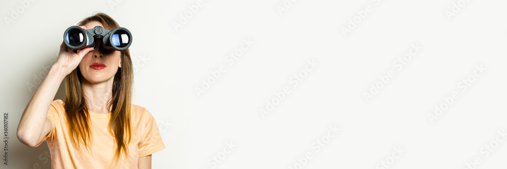 Young friendly girl looking through binoculars on a light background. Banner. Emotional face Young friendly girl looking through binoculars on a light background. Banner. Emotional face