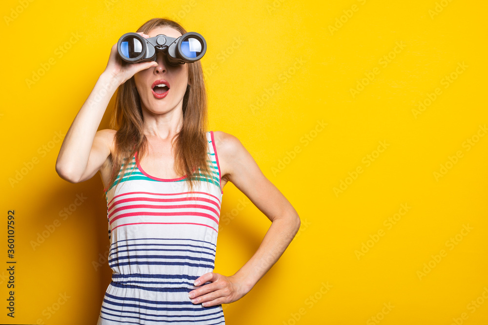 Young woman in a striped dress looks in shock with surprise in binoculars on a yellow background