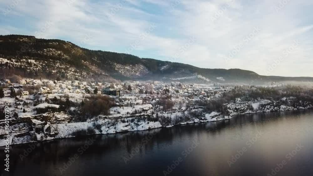 Scenic winter flight above still cold dark waters towards small snowy quaint village in Norway on sunny day, overhead drone approach Scenic winter flight above still cold dark waters towards small snowy quaint village in Norway on sunny day, overhead drone approach