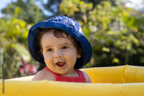 Bebe tomando sol com chapéu na cabeça, dentro de uma piscina infalível e brincando com a agua. Muito linda fazendo graças e sorrindo.