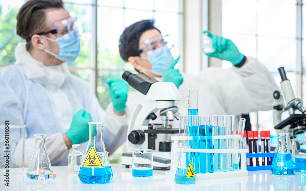 Lab equipment with blue liquid inside stand on the table with couple male scientist wearing protection suit holding vaccine for research at laboratory at the background. Lab equipment with blue liquid inside stand on the table with couple male scientist wearing protection suit holding vaccine for research at laboratory at the background.