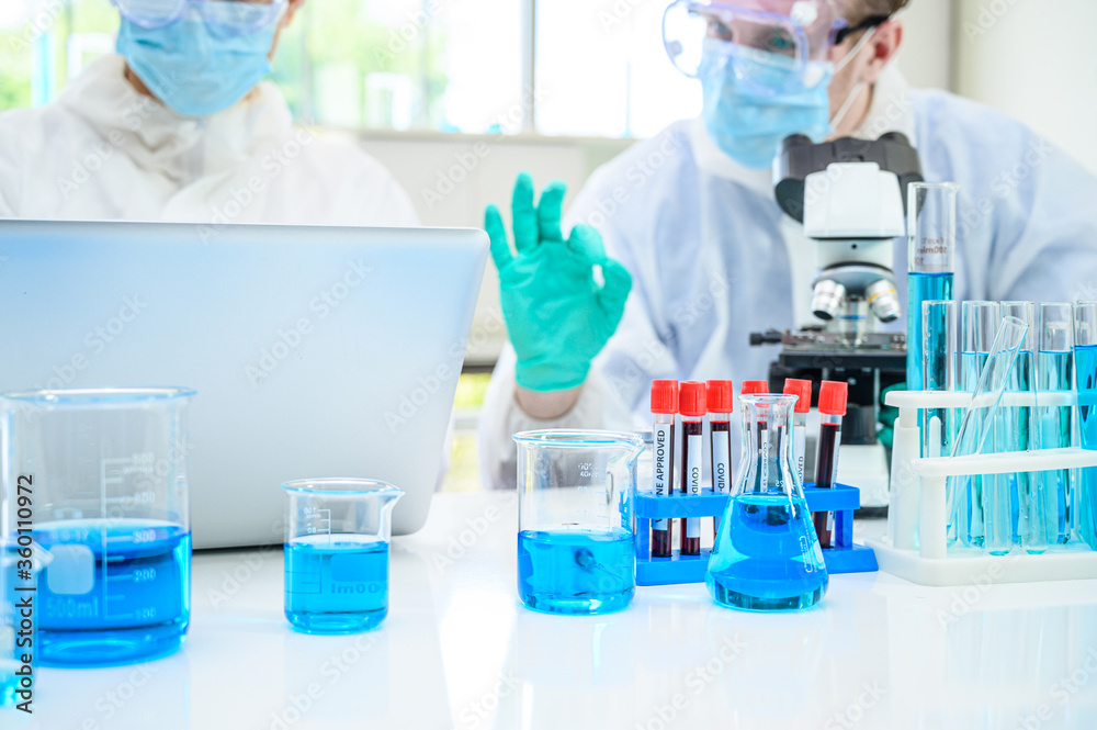Lab equipment with blue liquid inside stand on the table with couple male scientist wearing protection suit working at the background. Lab equipment with blue liquid inside stand on the table with couple male scientist wearing protection suit working at the background.